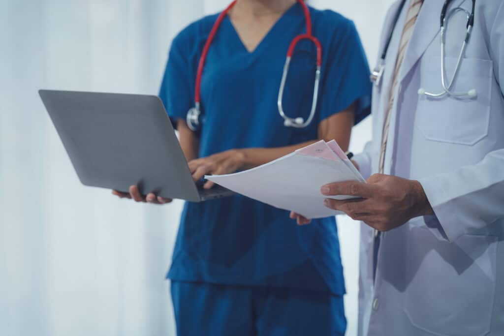 Professionnels de Santé: Consultation et Gestion des Dossiers Deux professionnels de santé. Infirmière en scrubs bleus avec stéthoscope utilise un ordinateur portable, un médecin en blouse lit des documents.