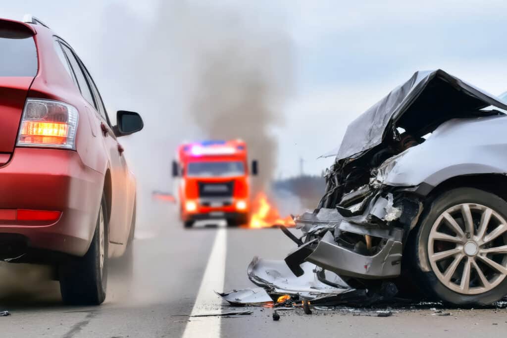 Accident de Voiture Grave : Incendie et Intervention des Pompiers Accident de voiture grave avec épave frontale en feu sur route, pompier et véhicule rouge en arrière-plan. Urgence routière.