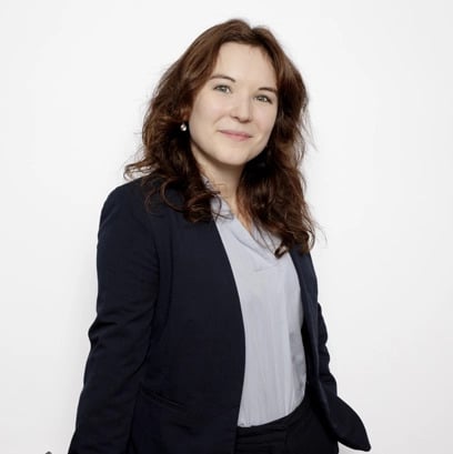 Portrait professionnel d'une femme souriante aux cheveux bruns, blazer bleu marine et chemise claire sur fond blanc.