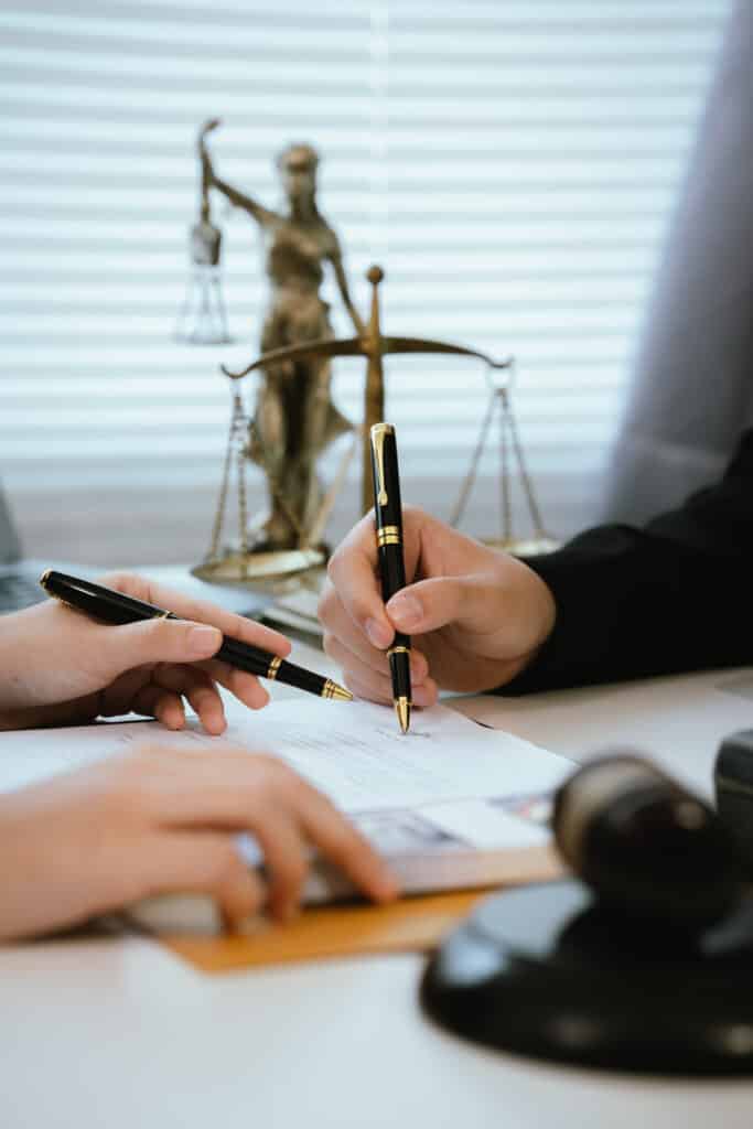 Signature Contrat Légal: Accord Officiel avec Avocat Deux mains signent un contrat avec des stylos noirs devant une statue de la Justice et des balances, symbole légal.