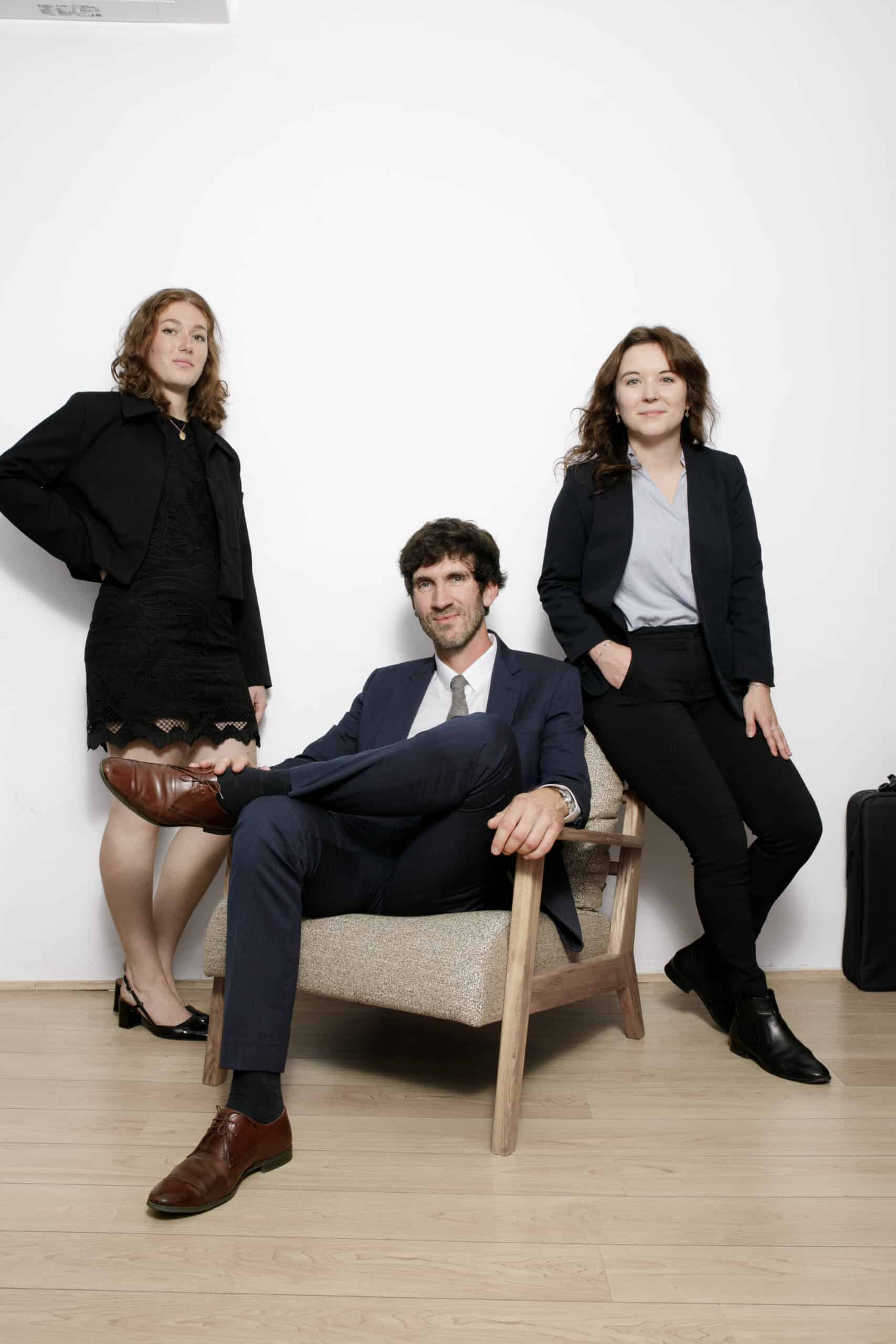 Portrait de trois professionnels : un homme en costume assis et deux femmes en blazer noir, posant avec assurance sur fond blanc uni.