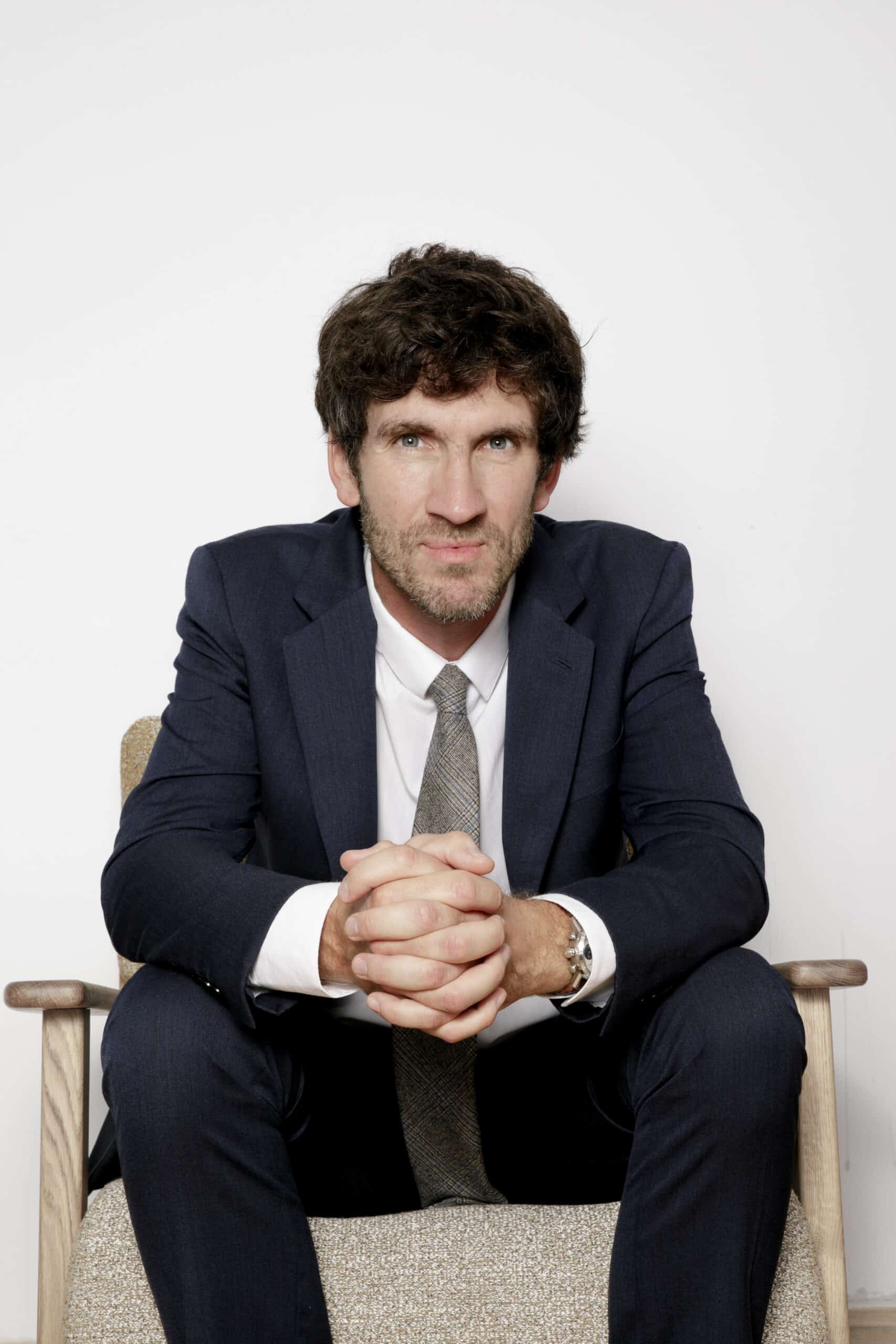 Homme professionnel en costume marine, chemise blanche et cravate. Assis sur une chaise bois, il croise les mains avec un regard intense.