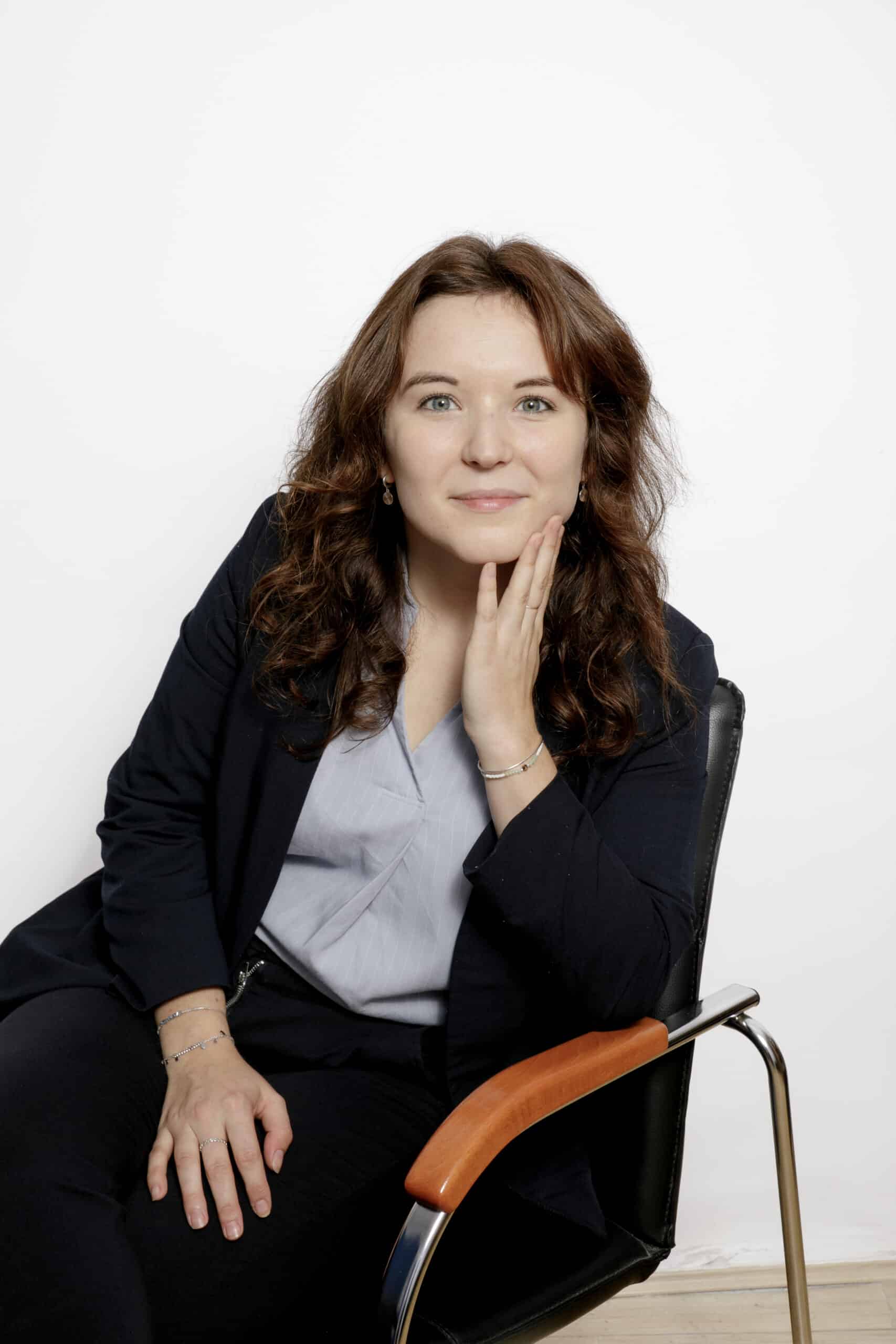 Portrait d'une jeune professionnelle souriante en blazer marine. Assise, elle repose pensivement sa main droite sur sa joue. Fond blanc.