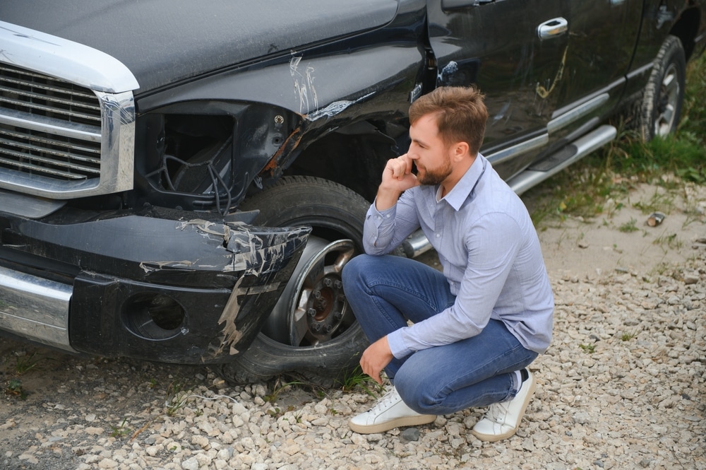 Homme inquiet au téléphone devant son pick-up noir accidenté avec phare brisé et pneu crevé sur du gravier.